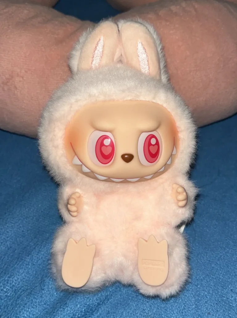 Close-up of a light pink Labubu plush toy, featuring its playful expression and fluffy bunny costume. The plush showcases large, heart-shaped eyes and is held against a soft blue background. The Pop Mart branding is visible on the bottom of the toy, emphasizing its authenticity