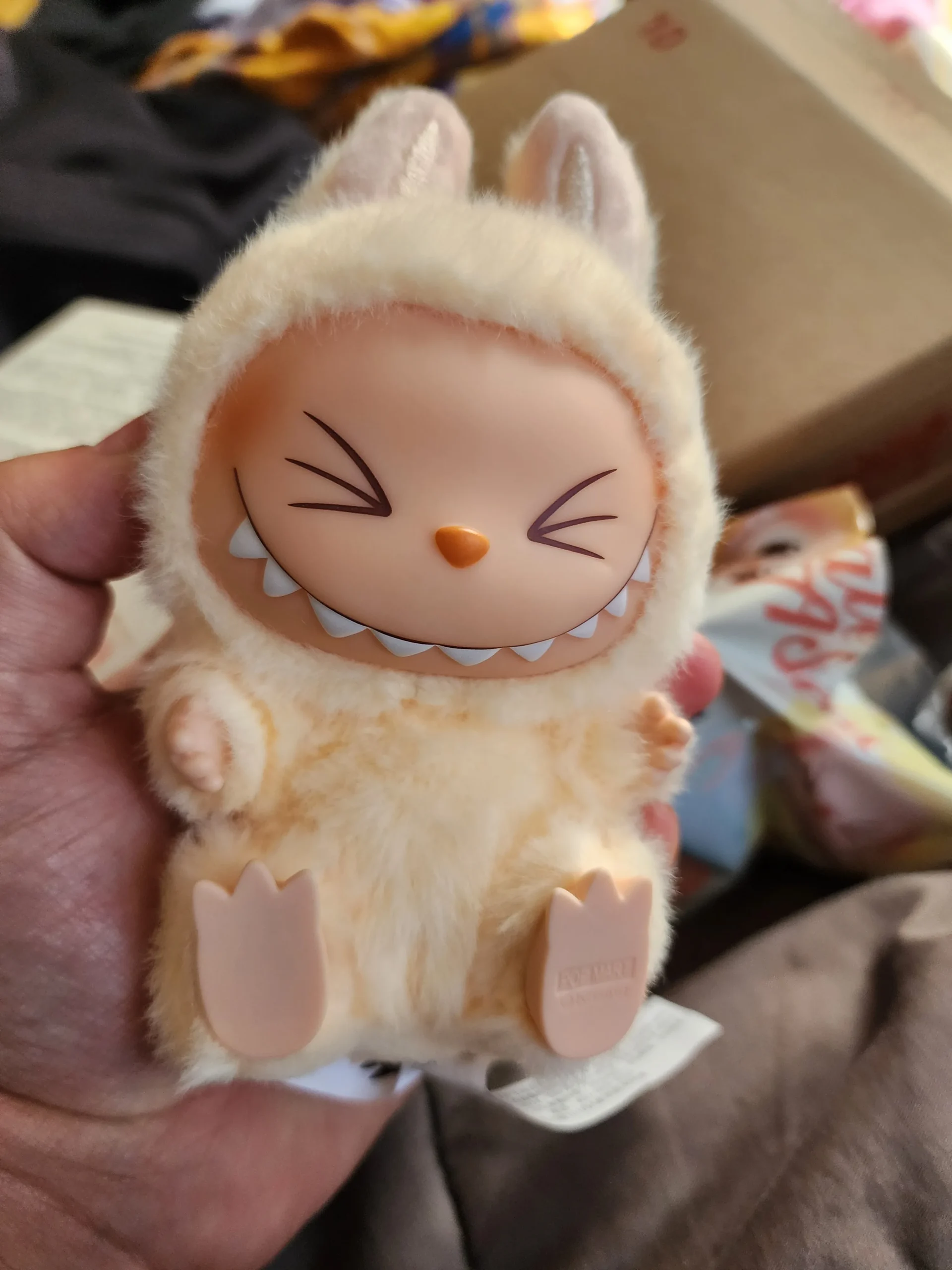 Close-up of a smiling Labubu plush toy wearing a soft, beige bunny costume. The toy has a wide, cheerful grin with exposed teeth, held in hand. The Pop Mart tag is visible at the base, and the background shows a cozy setting with a partially visible box