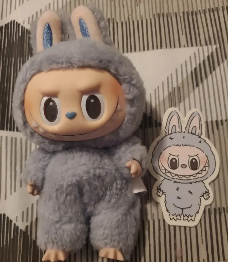 Close-up of a blue Labubu plush toy in a soft, fluffy bunny costume with large, expressive eyes and a mischievous grin. A matching Labubu sticker featuring the same character in a grey bunny costume is placed next to the plush. The background features a geometric patterned surface