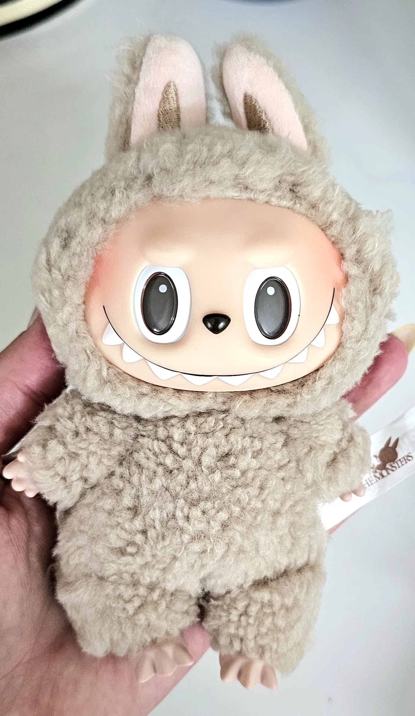 Close-up of a beige Labubu plush toy dressed in a fluffy bunny costume. The plush has a wide smile with exposed teeth and large expressive eyes. The toy is held in hand with the Pop Mart tag visible, showcasing its authenticity. The background is neutral, highlighting the plush's soft texture