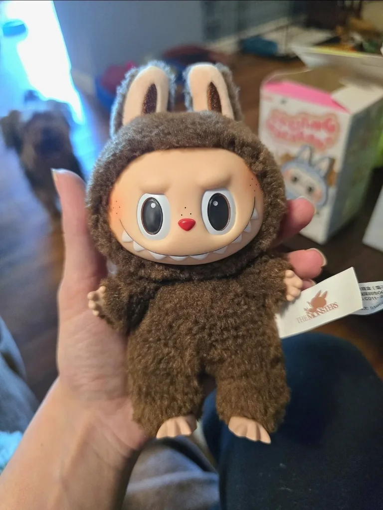 Close-up of a brown Labubu plush toy wearing a fluffy bunny costume with expressive eyes and a mischievous grin. The plush is held in hand, with the Pop Mart tag visible. In the background, a box and a blurred dog can be seen, adding a cozy home setting