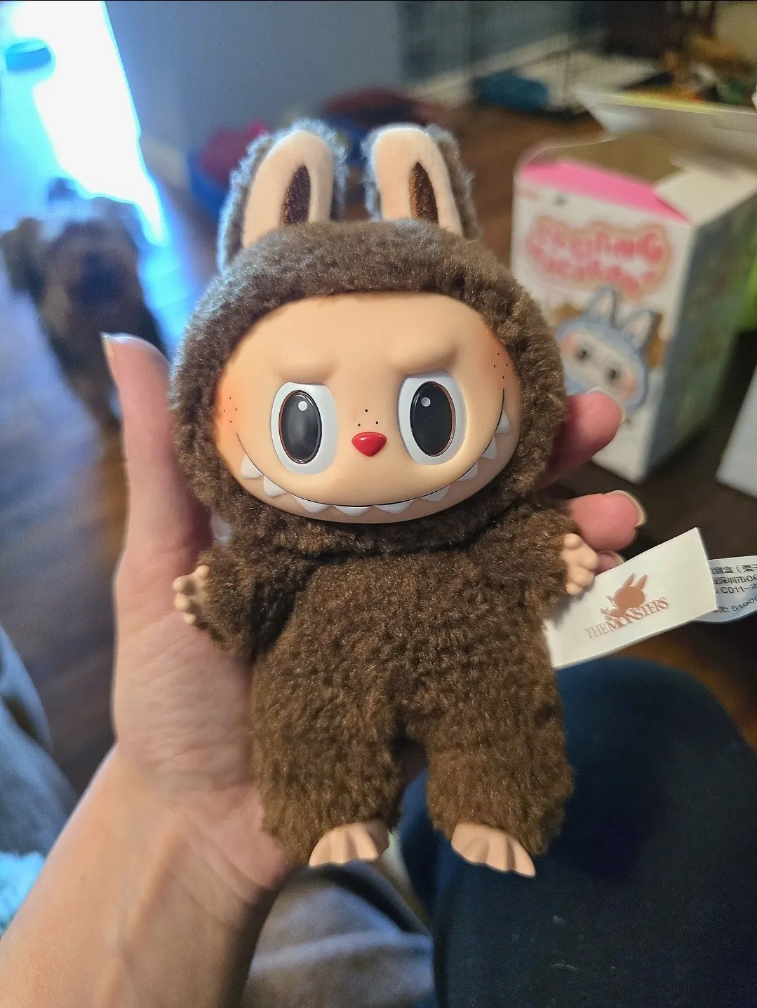 Close-up of a brown Labubu plush toy wearing a fluffy bunny costume with expressive eyes and a mischievous grin. The plush is held in hand, with the Pop Mart tag visible. In the background, a box and a blurred dog can be seen, adding a cozy home setting