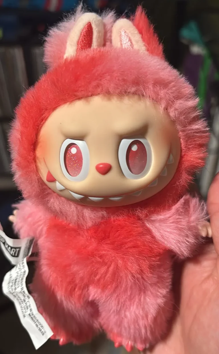 Close-up of a pink Labubu plush toy featuring its signature mischievous expression. The toy is dressed in soft, fluffy pink fur with matching bunny ears and a playful grin. The plush also displays the Pop Mart tag, highlighting its official authenticity