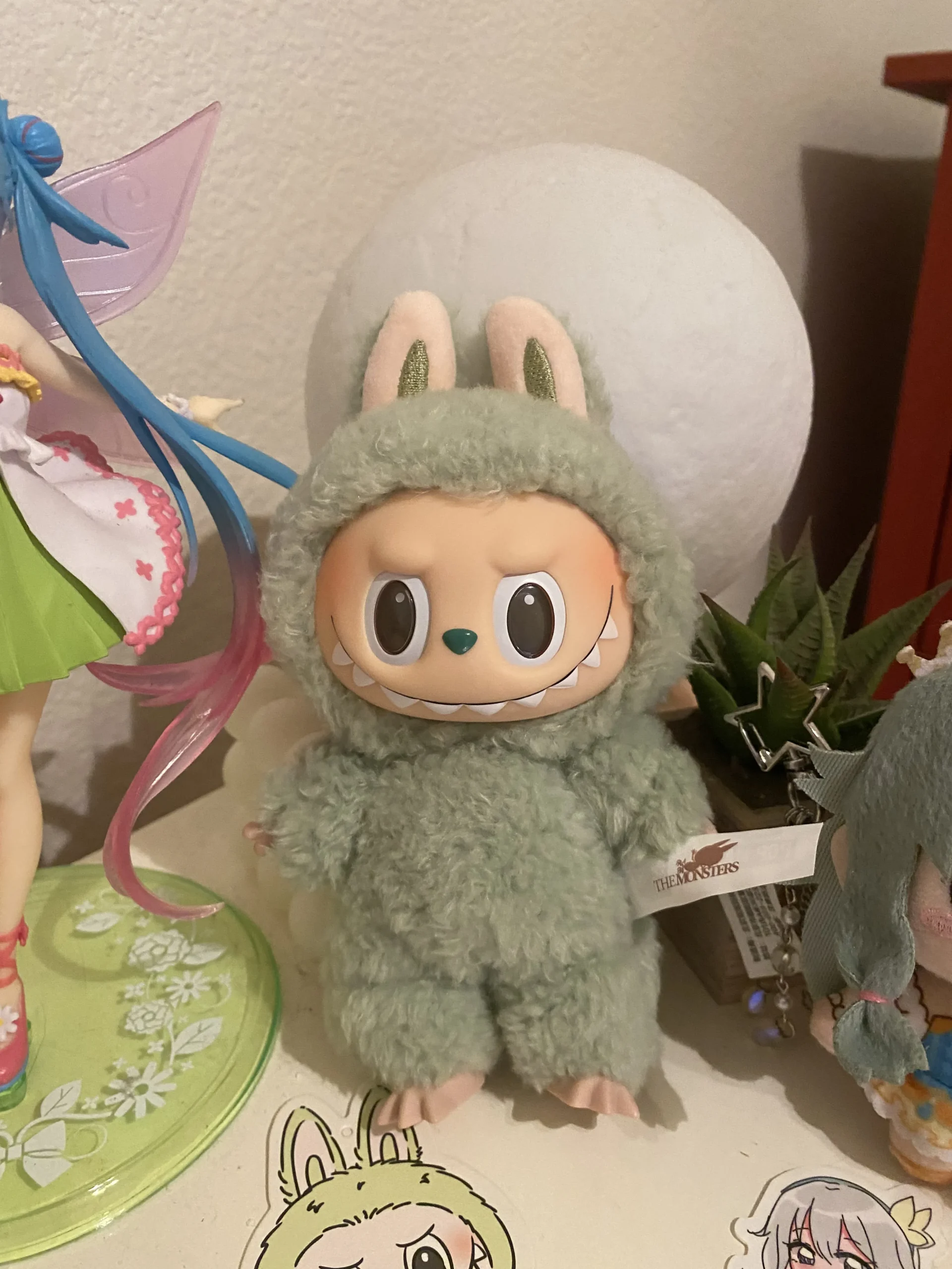 Labubu plush toy in a soft green bunny costume, featuring expressive eyes and a playful grin. The plush is placed on a shelf with various decorative items in the background, including figurines and plants. The Pop Mart tag is visible, ensuring its authenticity