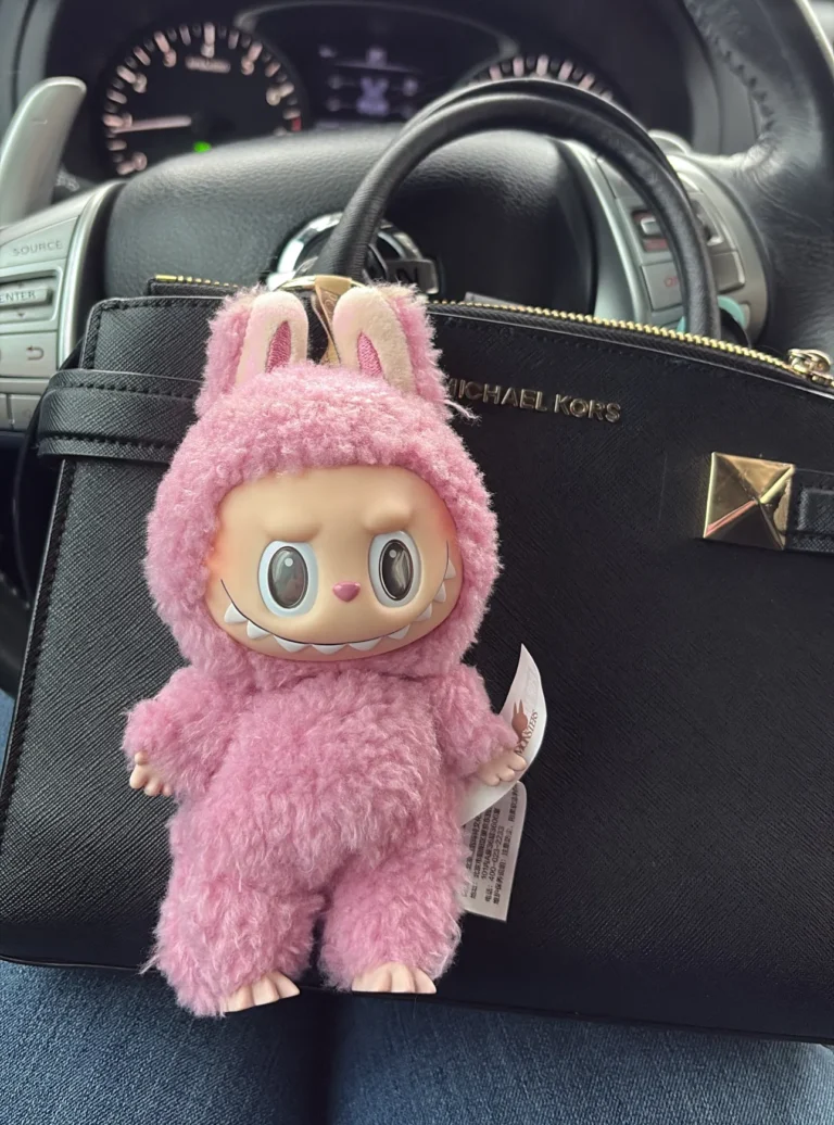 Pink Labubu plush toy wearing a fluffy bunny costume, with big eyes and a playful grin. The plush is placed on the seat next to a black Michael Kors bag inside a car. The Pop Mart tag is visible on the plush, showcasing its authenticity