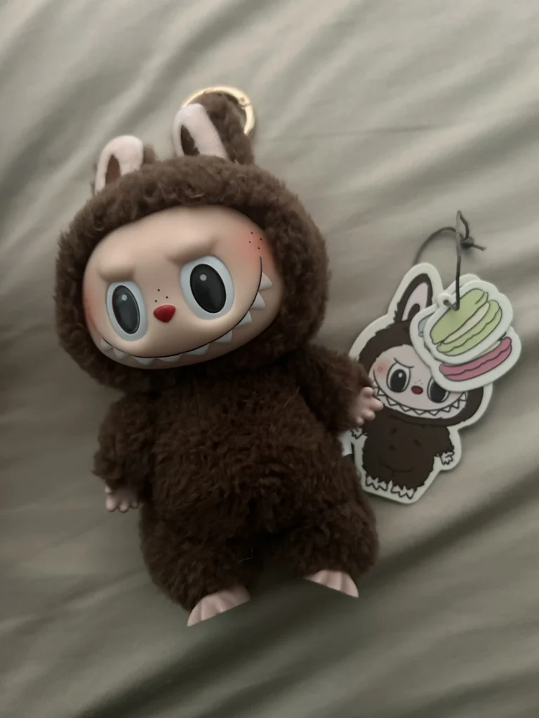 Brown Labubu plush toy in a fluffy bunny costume with a playful expression, holding a keychain. The plush is accompanied by a sticker featuring a cute Labubu design. The plush’s Pop Mart tag is visible, ensuring its authenticity, with a soft neutral-colored background