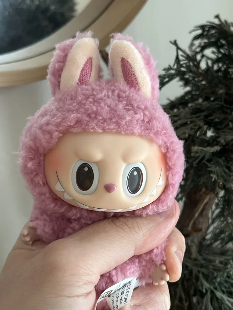 Close-up of a pink Labubu plush toy in a soft, fluffy bunny costume with expressive eyes and a playful grin. The plush is held in hand, with the Pop Mart tag visible on its side. A mirror and a decorative plant provide a cozy background