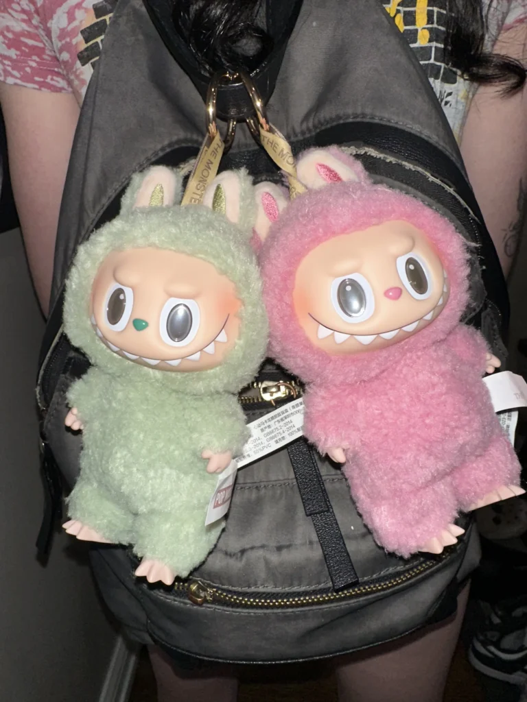 Two Labubu plush toys in soft bunny costumes, one in green and the other in pink, hanging from a backpack with visible Pop Mart tags. The plush toys have playful expressions, and the background features part of the backpack and a casual outfit