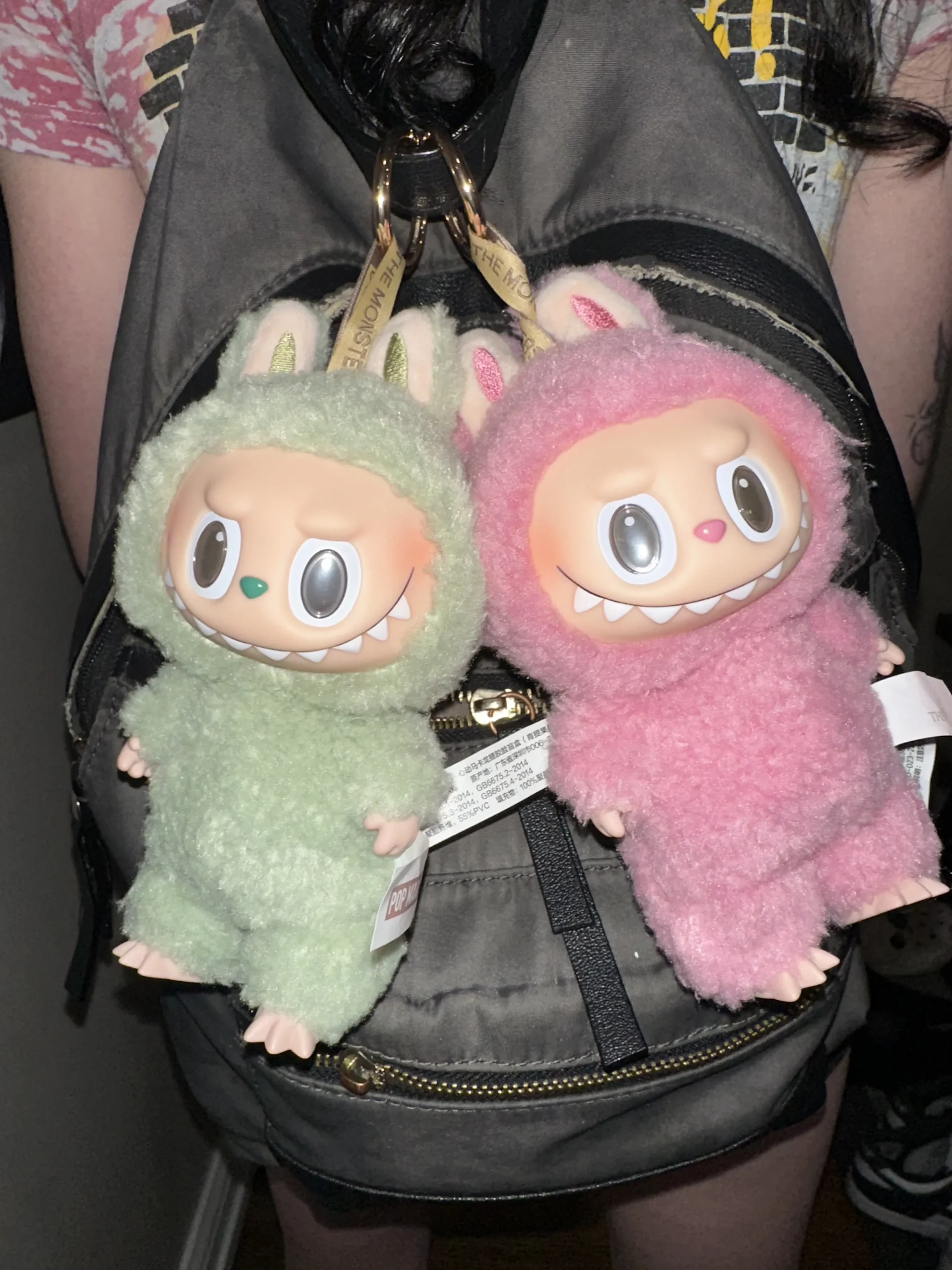 Two Labubu plush toys in soft bunny costumes, one in green and the other in pink, hanging from a backpack with visible Pop Mart tags. The plush toys have playful expressions, and the background features part of the backpack and a casual outfit