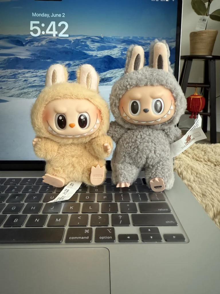 Two Labubu plush toys, one in beige and the other in grey, both wearing soft bunny costumes with mischievous expressions. They are placed on a laptop keyboard with visible Pop Mart tags, and the background features a scenic mountain wallpaper and a plant
