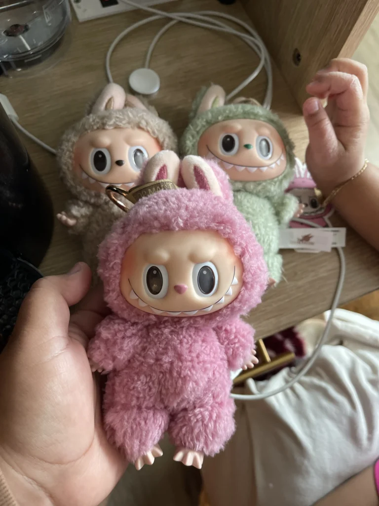 A collection of three Labubu plush toys, each in a fluffy bunny costume in different colors (pink, green, and beige), with their signature playful expressions. One plush is being held in hand while the others are positioned on a wooden surface, with visible Pop Mart tags. A child's hand is seen in the background, adding to the cozy home setting