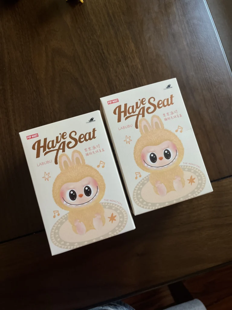 Two 'Have A Seat' Labubu collectible boxes from Pop Mart, each featuring the adorable Labubu character in a fluffy bunny costume, sitting in a playful pose. The boxes have musical notes and a soft pastel color palette, with the Pop Mart and The Monsters branding visible