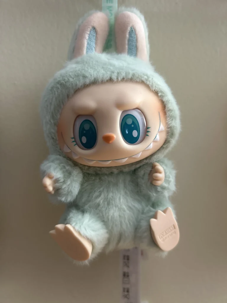Close-up of a light blue Labubu plush toy, wearing a soft bunny costume with pink ears. The plush features Labubu’s playful yet mischievous expression and is displayed with the Pop Mart tag visible at the base. The toy is hanging against a neutral background