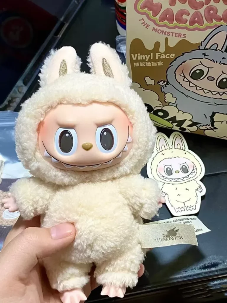 A beige Labubu plush toy in a fluffy bunny costume, with a mischievous expression and large eyes, held in hand. The plush is accompanied by a sticker featuring the same design and a box labeled 'Vinyl Face.' The Pop Mart tag is visible, showcasing the toy’s authenticity