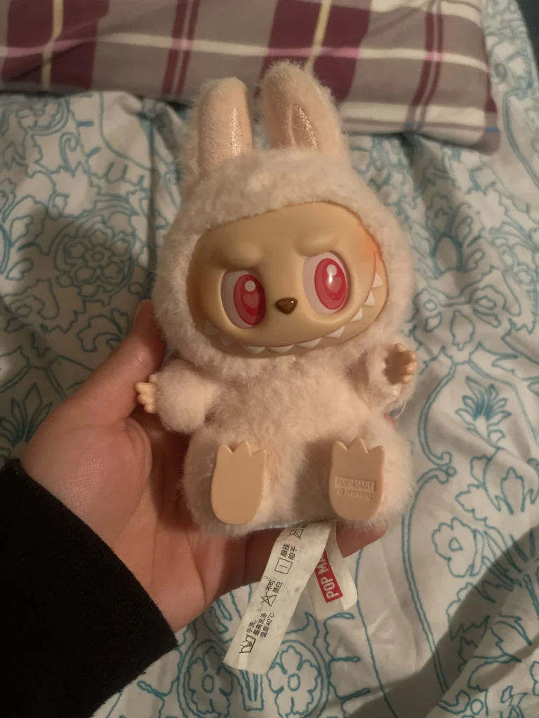 Close-up of a soft pink Labubu plush toy in a bunny costume, with large red eyes and a mischievous grin. The plush is held in hand, and the Pop Mart tag is visible at the bottom. The background shows a cozy bed with floral sheets, creating a relaxed vibe