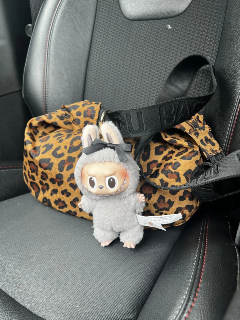 Gray Labubu plush toy in a fluffy bunny costume, with an expressive face and visible Pop Mart tag, placed on a leopard print bag in a car. The plush has a playful, mischievous look, and the background features the car's interior details