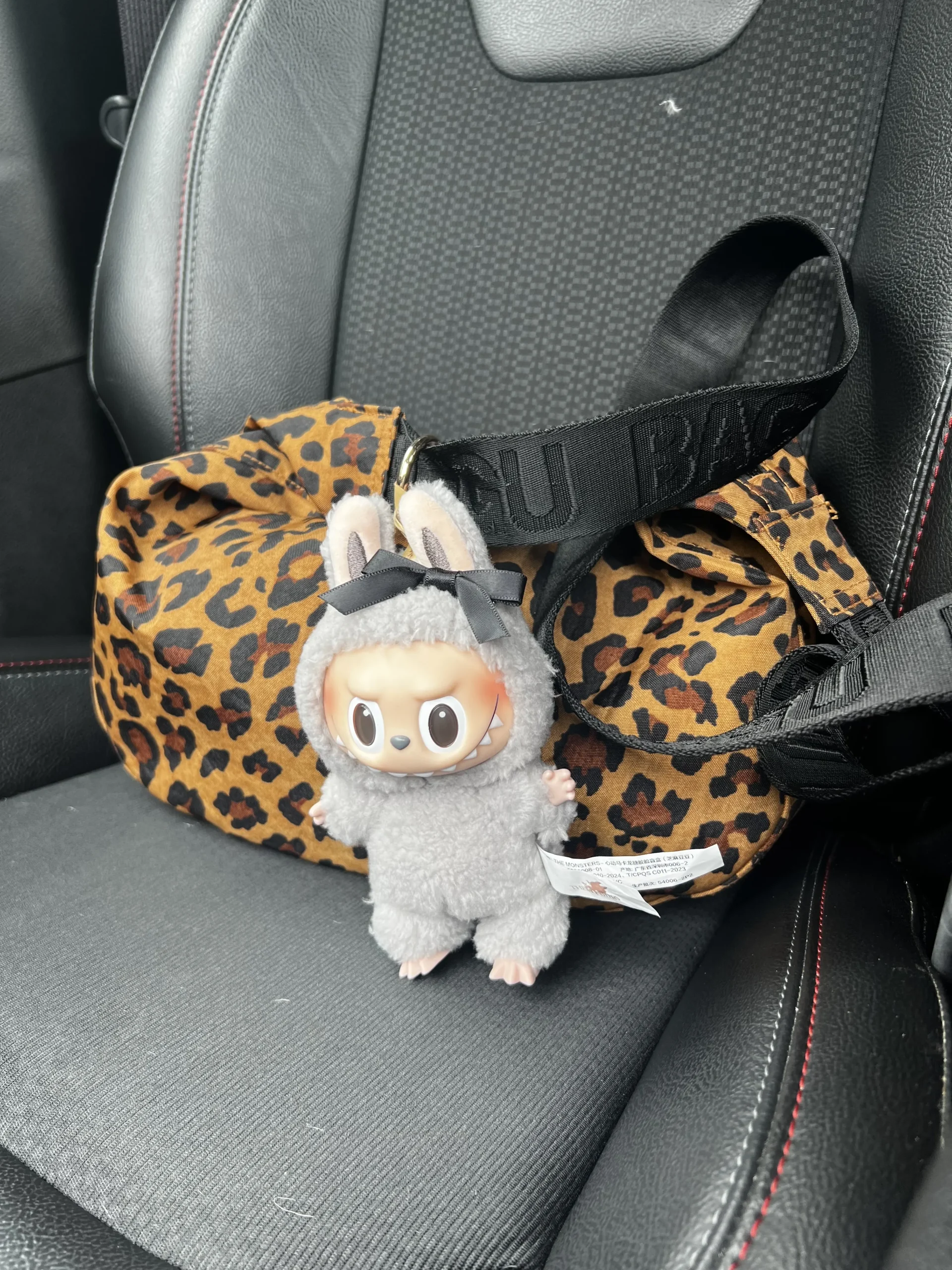 Gray Labubu plush toy in a fluffy bunny costume, with an expressive face and visible Pop Mart tag, placed on a leopard print bag in a car. The plush has a playful, mischievous look, and the background features the car's interior details